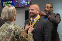 Col. Michelle M. Williams, U.S. Army Financial Management Command commander, left, and Retired Command Sgt. Maj. Gennaro Penn, U.S. Army Finance Corps Association president, right, present Mark R. W. Orders-Woempner, USAFMCOM public affairs officer, with a U.S. Army Finance Corps’ Honorary Member of the Regiment award on behalf of Col. Robert Le'iato, U.S. Army Finance and Comptroller School commandant and Chief of the Finance Corps, at the Maj. Gen. Emmett J. Bean Federal Center in Indianapolis Jan. 12, 2026. The Finance Corps designated the first Honorary Member of the Regiment on July 5, 1988, to honor those who made significant professional contributions or have brought great credit to the Finance Corps through special acts or accomplishments. (U.S. Army photo by Maj. Terrell Murdock)