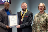Col. Michelle M. Williams, U.S. Army Financial Management Command commander, right, and Retired Command Sgt. Maj. Gennaro Penn, U.S. Army Finance Corps Association president, left, present Mark R. W. Orders-Woempner, USAFMCOM public affairs officer, with a U.S. Army Finance Corps’ Honorary Member of the Regiment award on behalf of Col. Robert Le'iato, U.S. Army Finance and Comptroller School commandant and Chief of the Finance Corps, at the Maj. Gen. Emmett J. Bean Federal Center in Indianapolis Jan. 12, 2026. The Finance Corps designated the first Honorary Member of the Regiment on July 5, 1988, to honor those who made significant professional contributions or have brought great credit to the Finance Corps through special acts or accomplishments. (U.S. Army photo by Maj. Terrell Murdock)