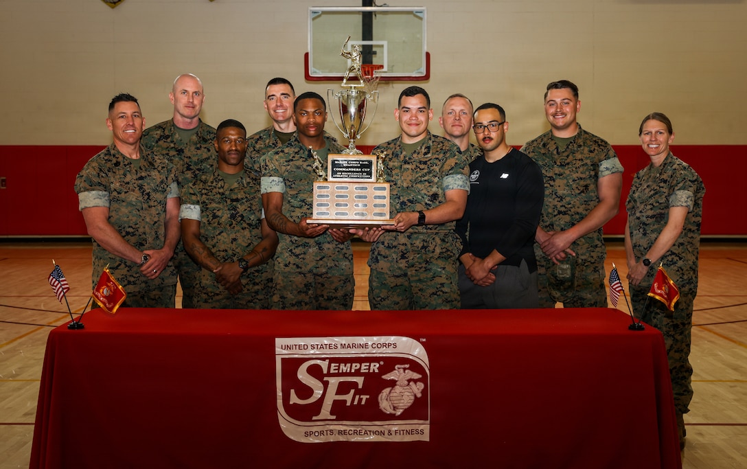 U.S. Marines with Security Battalion pose for a photo with U.S. Marine Corps Col. Jenny Colegate, the commanding officer of Marine Corps Base Quantico, after being present the Marine Corps Base Quantico Commander’s Cup Trophy at the Barber Physical Activity Center on MCB Quantico, Virginia, April 8, 2026. The MCB Quantico Commander’s Cup brings units from across the base together to compete in a variety of sports, all vying to come out on top. Security Battalion accumulated 130 Commander’s Cup points, earning first place overall. The battalion placed second in soccer, participated in softball, and won the championship in flag football. (U.S. Marine Corps photo by Lance Cpl. Jeffery Stevens)