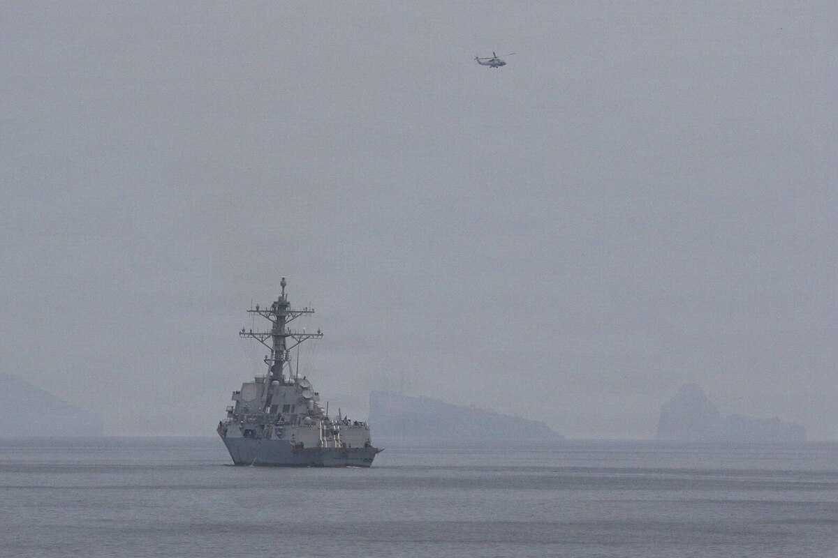 A large military ship sails in the water as a military helicopter flies overhead.