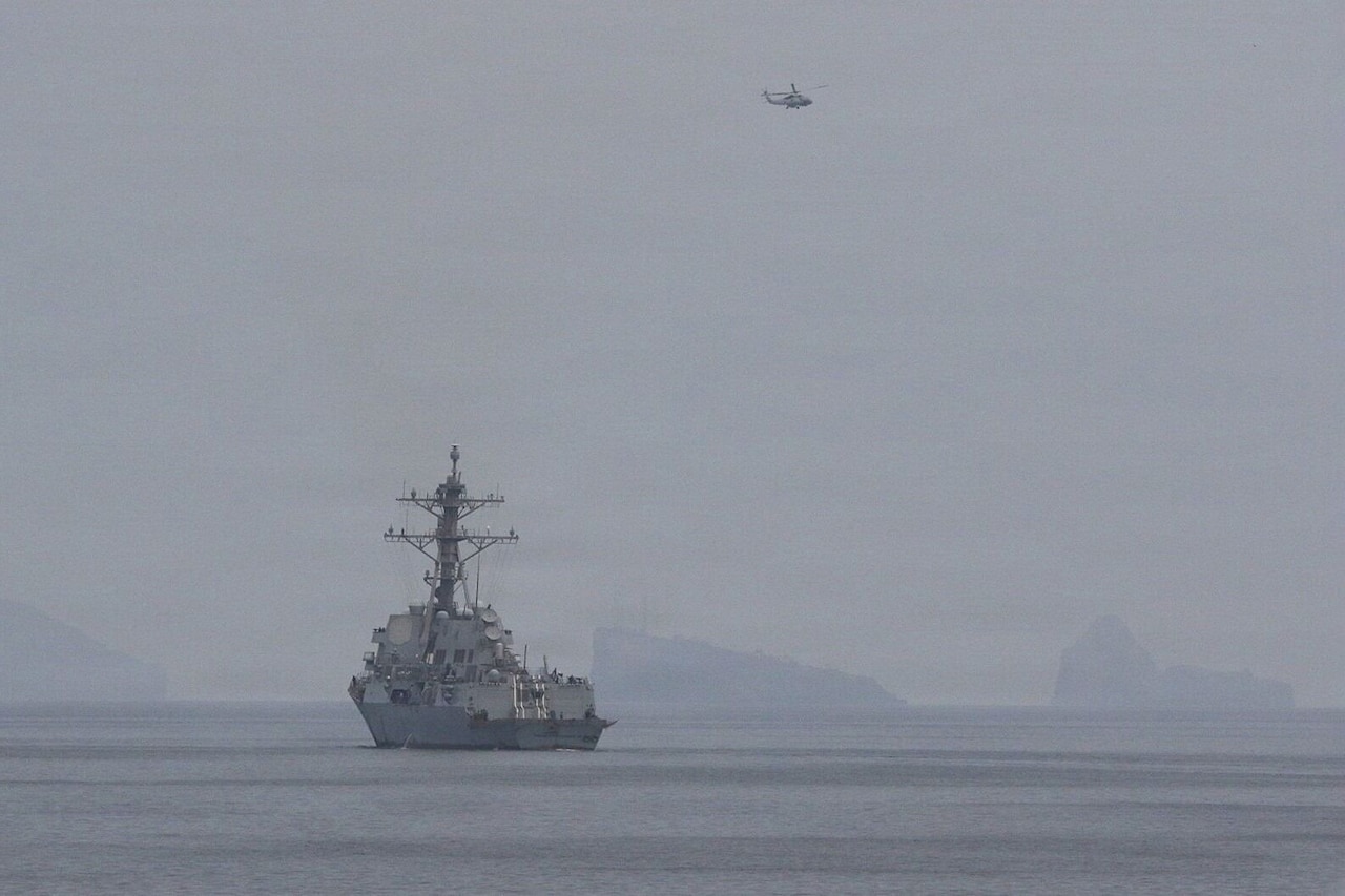 A large military ship sails in the water as a military helicopter flies overhead.