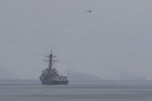 A large military ship sails in the water as a military helicopter flies overhead.