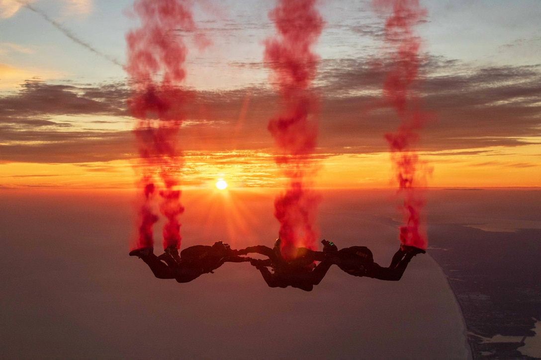 Four soldiers hold hands while descending in a sunlit sky with clouds of red smoke coming from their feet.