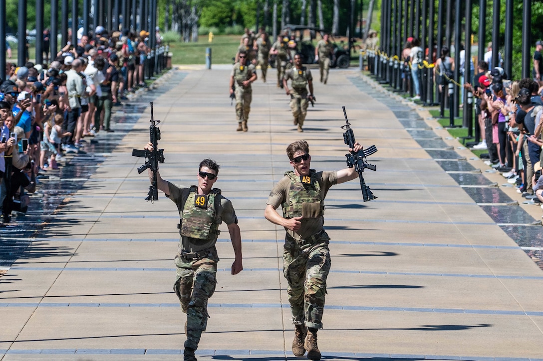 Spectators applaud and record as two soldiers in tactical gear hold weapons in the air while crossing the finish line.