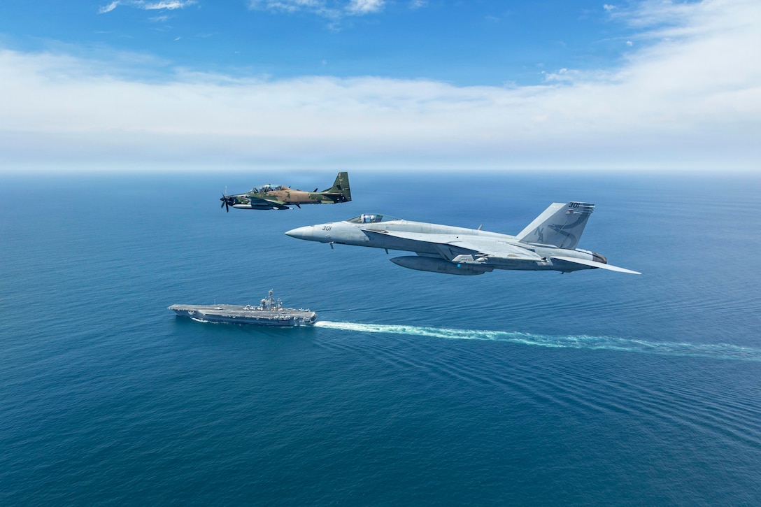 Two aircraft fly next to each other in a blue sky above a ship at sea.