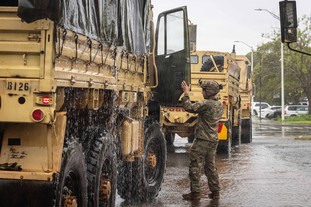 A soldier in uniform guides the driver of a military vehicle through the open passenger door as it rains.