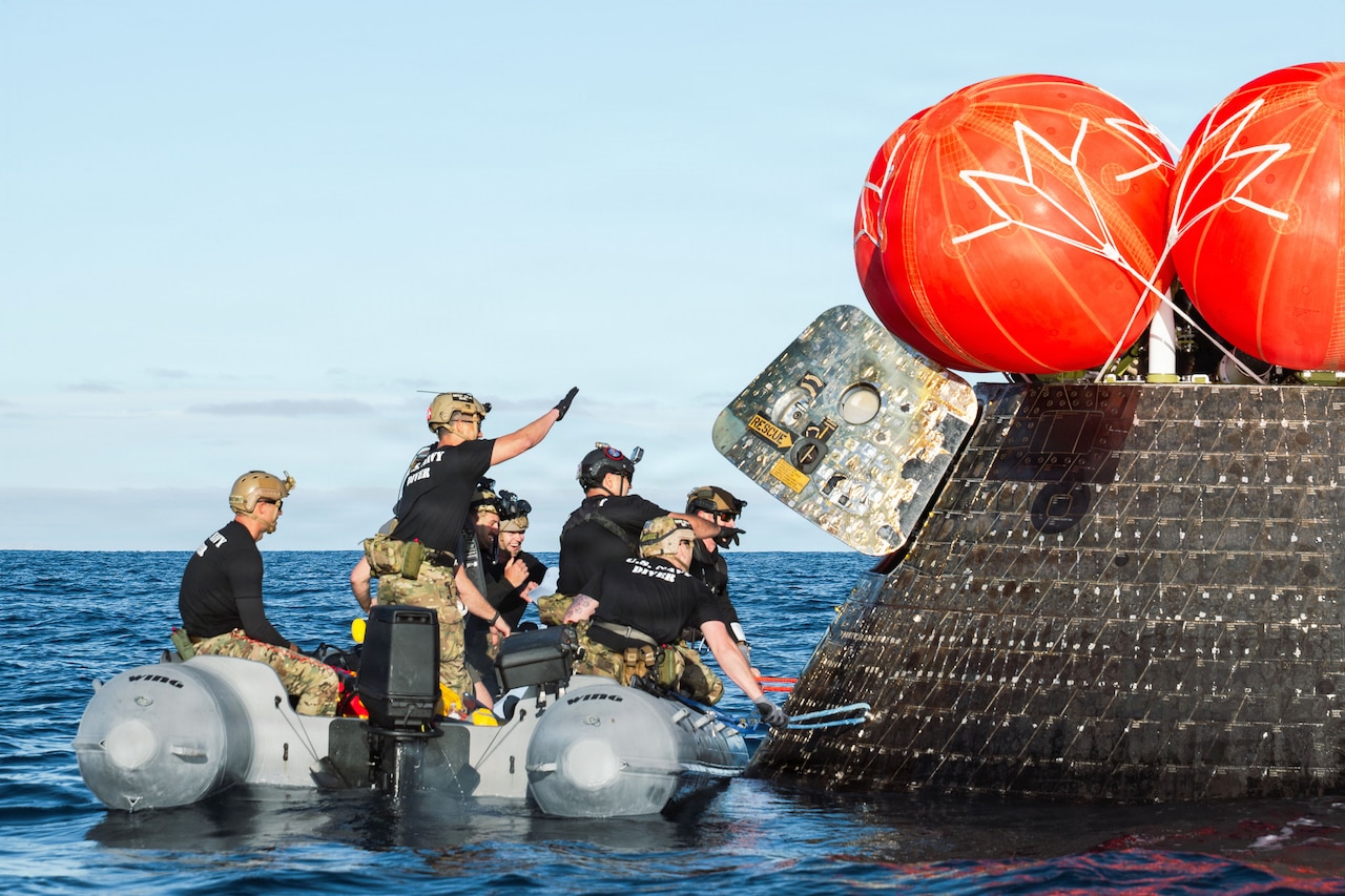 Sailors aboard a small boat approach a spacecraft in a body of water during the day.