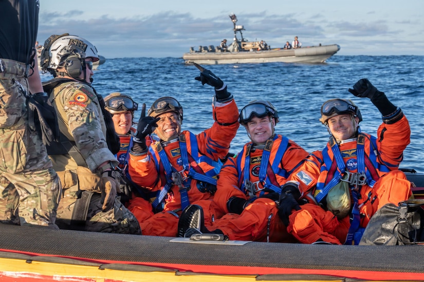 Four people in NASA spacesuits pose for a photo while sitting on a small vessel in the ocean that is manned by two people in military camouflage uniforms.