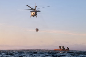 Two people are hoisted up a line into a military helicopter that is hovering over the ocean. Four other people are floating on a raft below the helicopter.