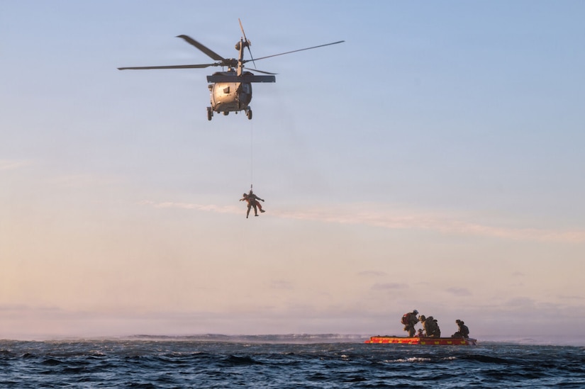 Two people are hoisted up a line into a military helicopter that is hovering over the ocean. Four other people are floating on a raft below the helicopter.
