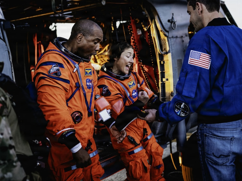 A man and a woman in NASA spacesuits look excited as a man in a flight suit gives them Navy command hats. The two astronauts are standing next to a military helicopter that is parked on the ship.