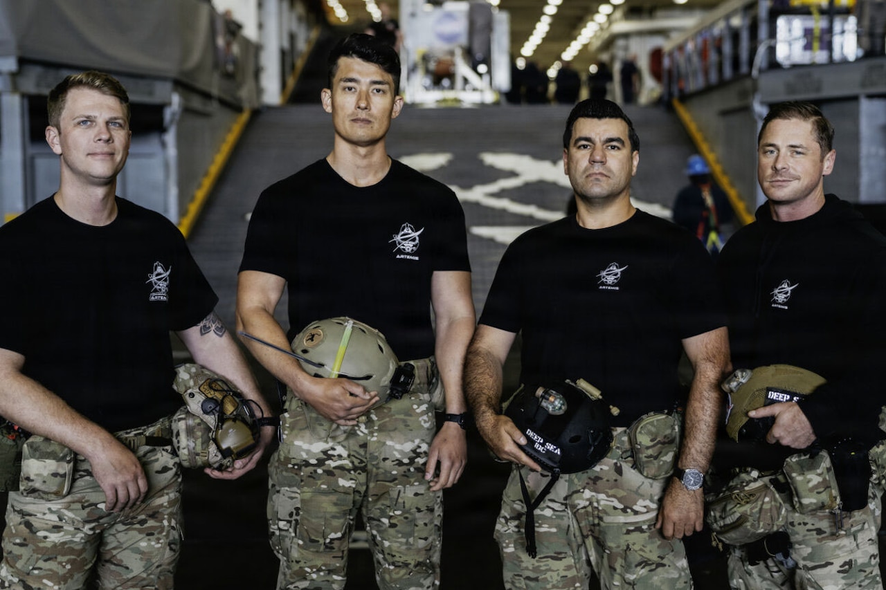 Four men in military uniforms pose for a photo inside a military transport ship sailing in the ocean.