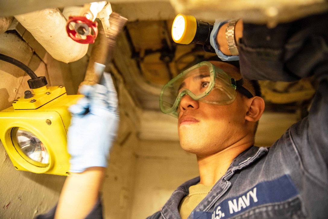 A sailor wearing goggles uses a flashlight to perform maintenance on a ship.