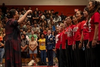 A woman conducts a student choir in the foreground while an Airman stands at attention at the back end of the line of students.