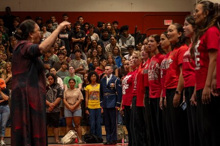 A woman conducts a student choir in the foreground while an Airman stands at attention at the back end of the line of students.