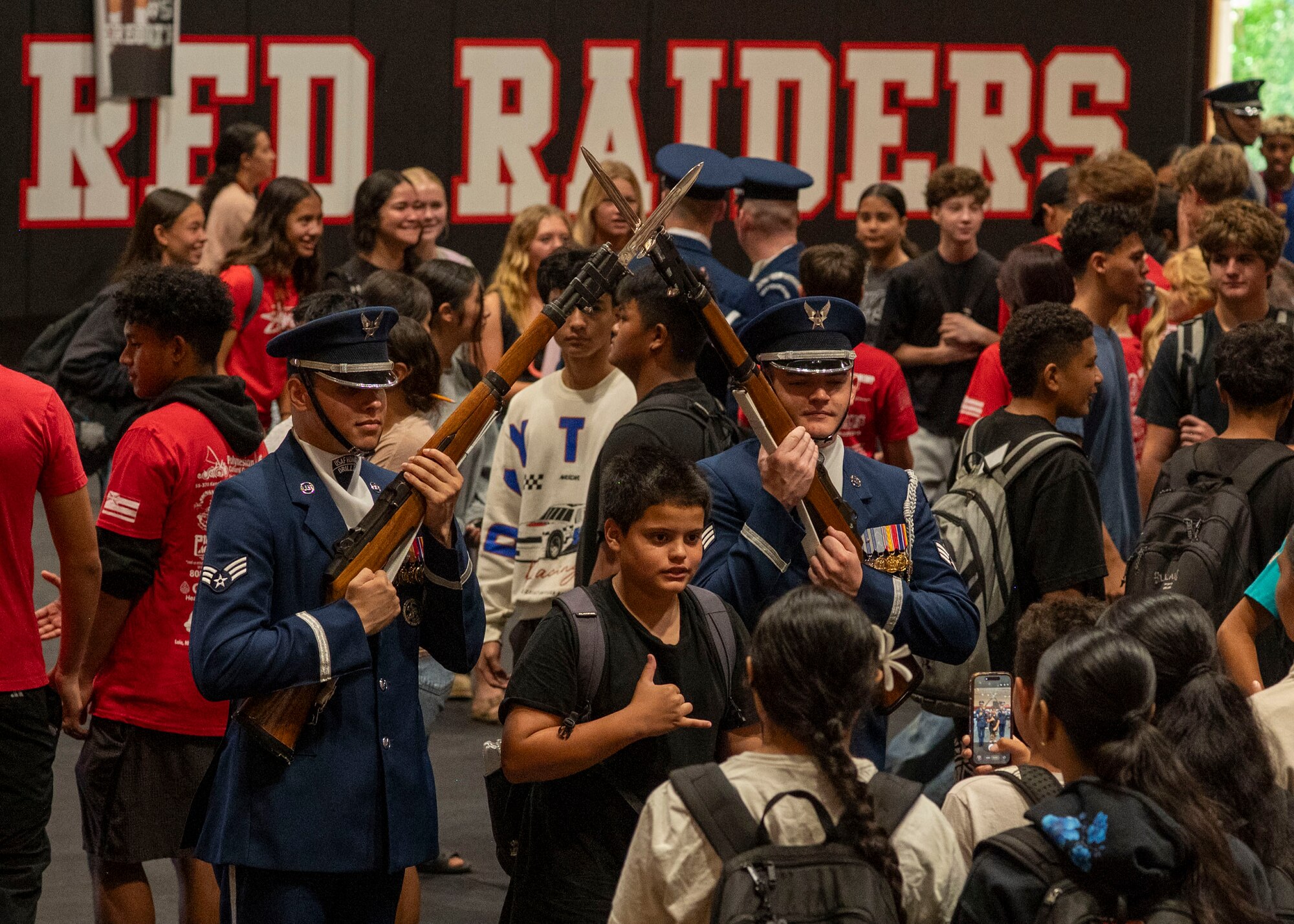 A student stands between two Air Force Honor Guard Drill Team members for a photo, showing a shaka hand sign.