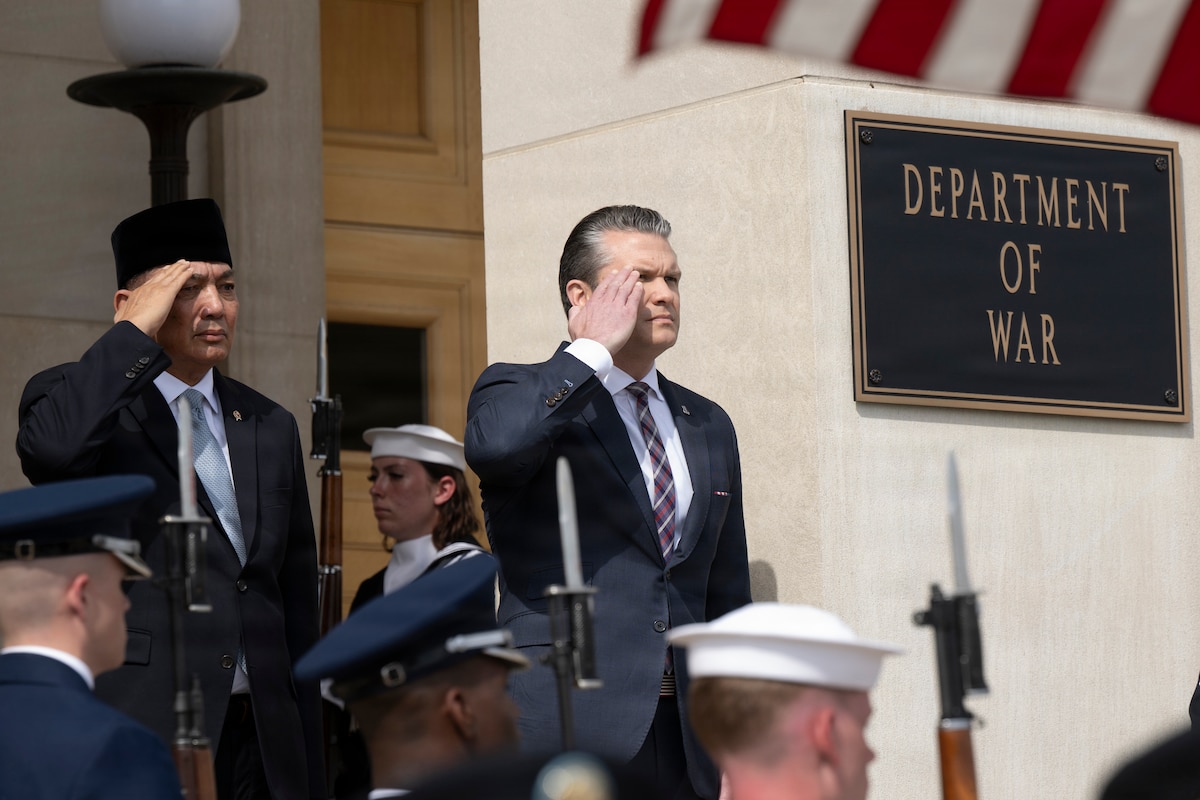 Two people in business attire salute as they stand outside during daytime, with service members standing at attention nearby.