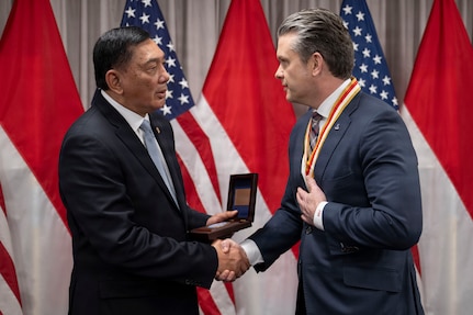 Two people in business attire shake hands as they stand indoors, with flags and a curtain in the background.