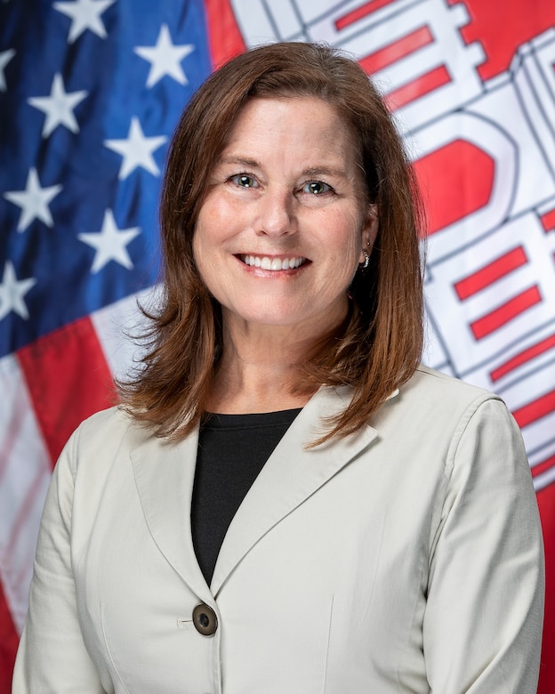 A woman in a beige suit jacket with brown shoulder length hair in front of the U.S. flag and the USACE flag.