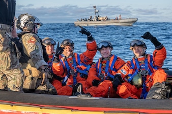Four people in NASA spacesuits pose for a photo while sitting on a small vessel in the ocean that is manned by two people in military camouflage uniforms.
