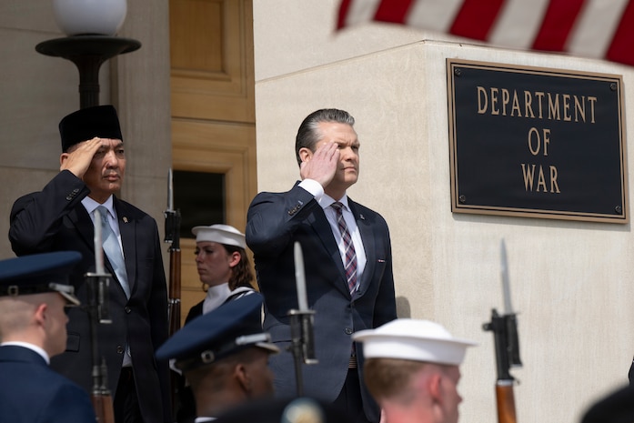Two people in business attire salute as they stand outside during daytime, with service members standing at attention nearby.