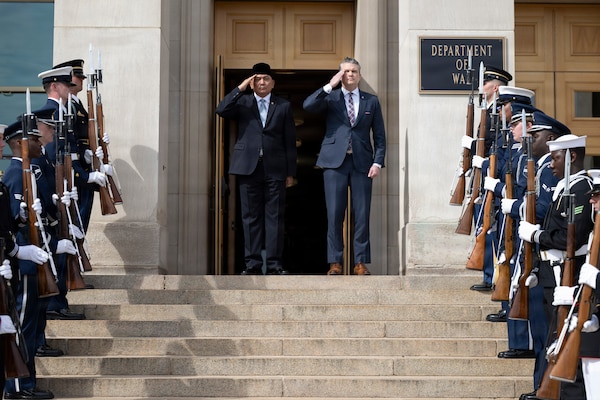 Two people in business attire salute as they stand outside during daytime, with service members standing at attention nearby.