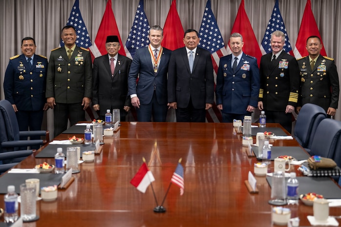 Three people in business attire and five people in formal military uniforms stand at the end of a table indoors, with flags and a curtain in the background.
