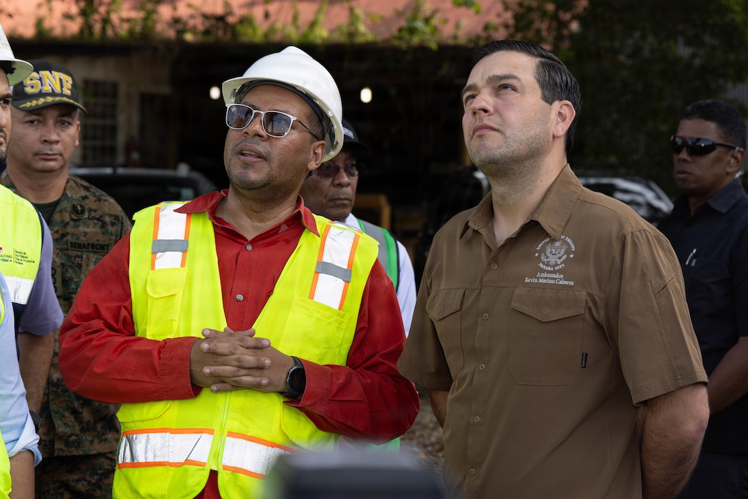 A man in a reflective vest and hard hat and a man in a brown shirt look up during a discussion.