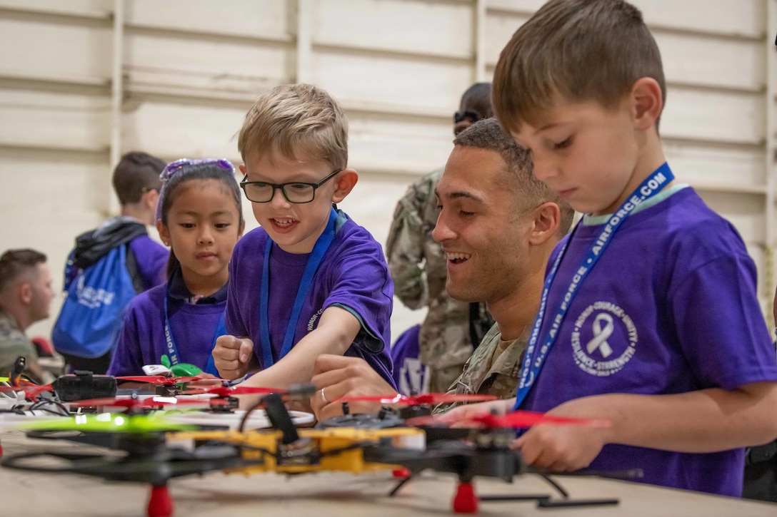 A service member kneels next to three kids in purple shirts as they touch drones sitting on a table.
