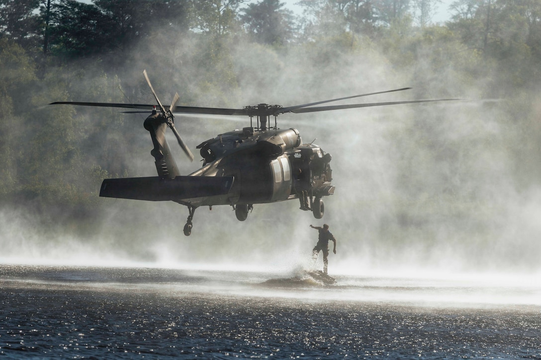 A soldier jumps into a pond from a helicopter hovering above during the day.