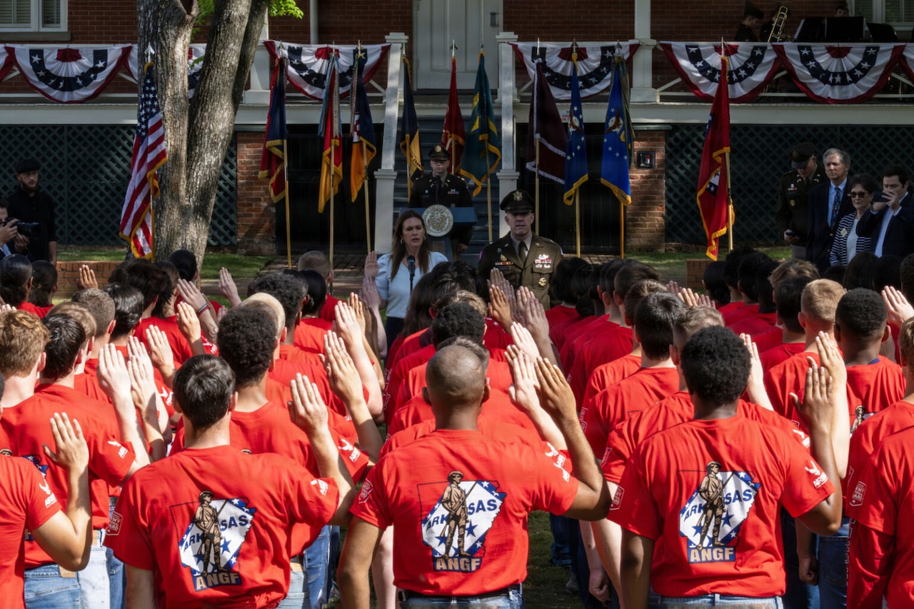 More than a dozen people wearing red T-shirts raise their right hand to take an oath of enlistment outside in front of a man wearing a formal military uniform and a woman in business attire; Behind them are an American flag and various other flags.