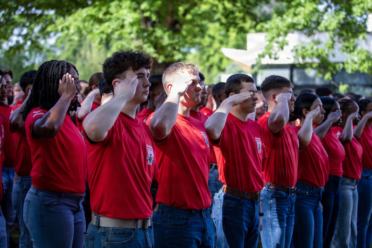 More than a dozen people wearing red T-shirts render a salute outside under a tree.