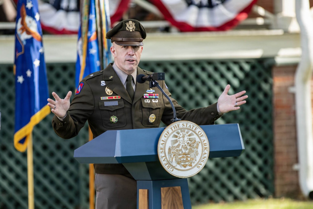 A man in a formal military uniform stands at a lectern with his arms outstretched, talking to an off-camera audience outside.