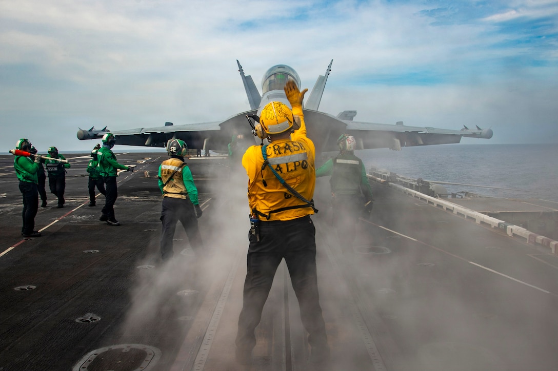 People on the deck of an aircraft carrier give hand signals to someone in an aircraft that is facing them, as smoke hangs in the air.