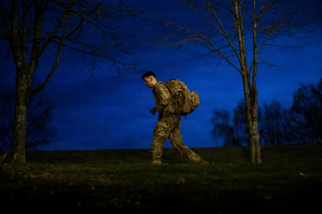 A soldier wearing a camouflage uniform and backpack looks ahead at the ground as he walks on grass, with a dark blue sky and a few trees in the background.