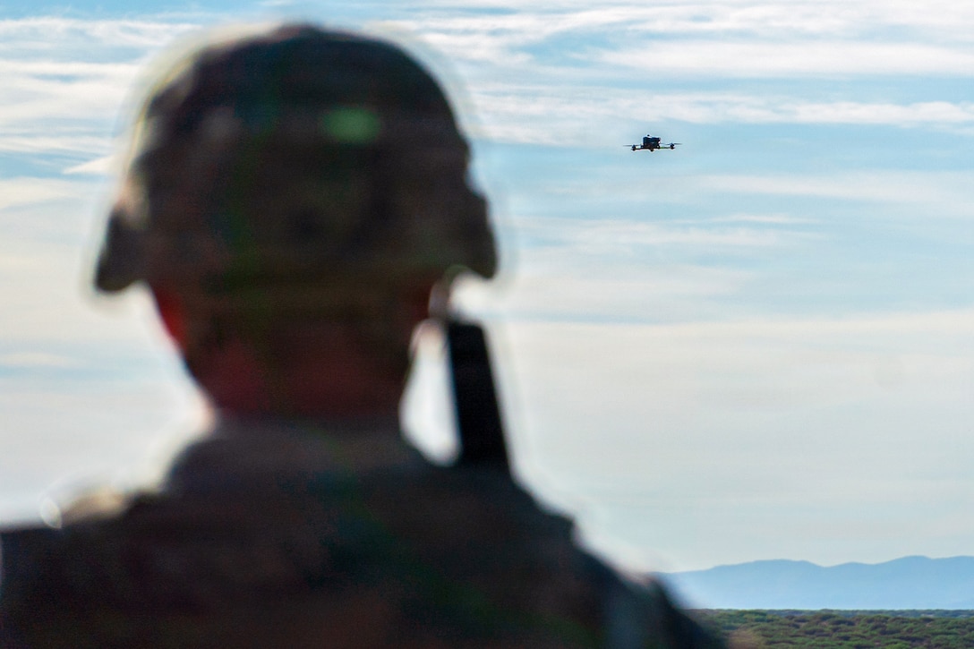 The back of a soldier wearing a helmet is seen and a drone is high in the light blue sky in the distance.