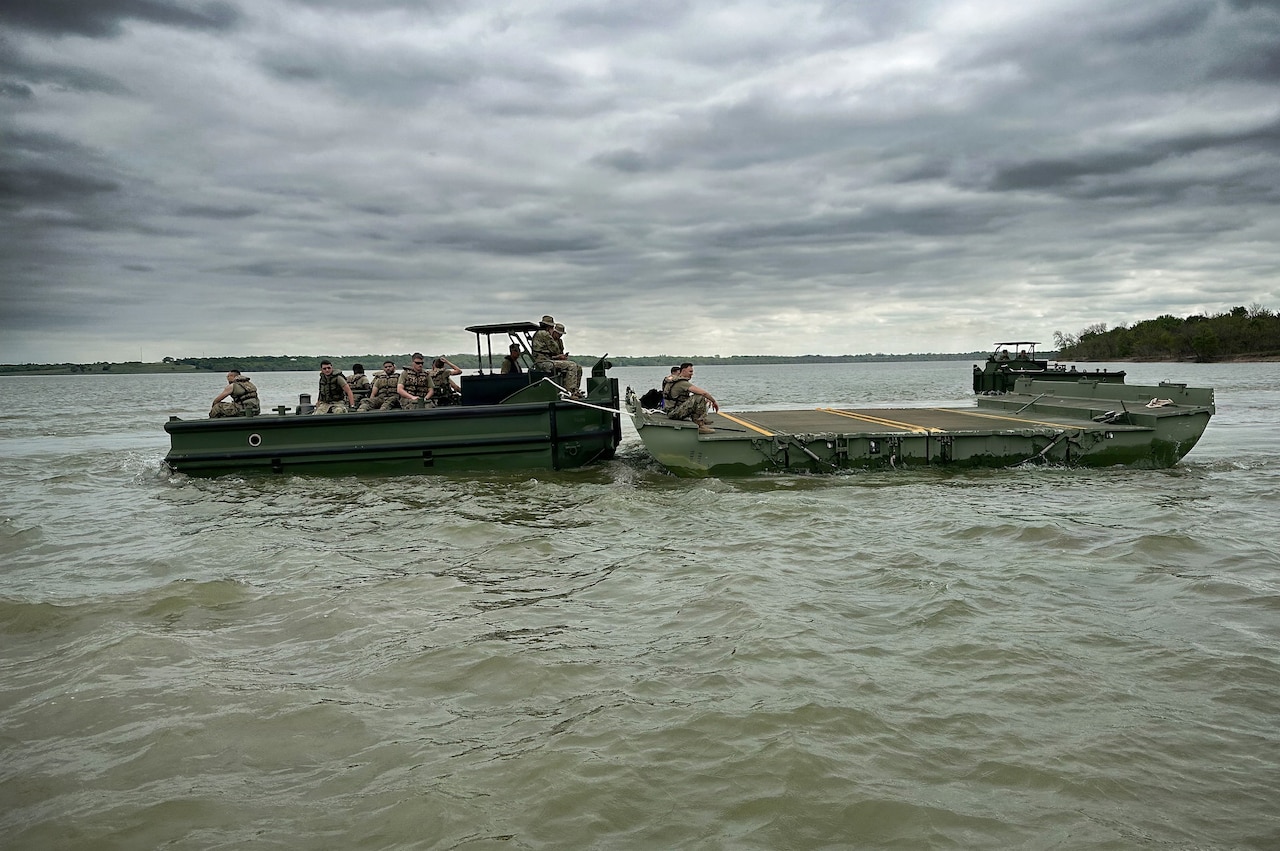 A dozen people in camouflage military uniforms and life jackets use a boat to maneuver a floating bridge in the water.