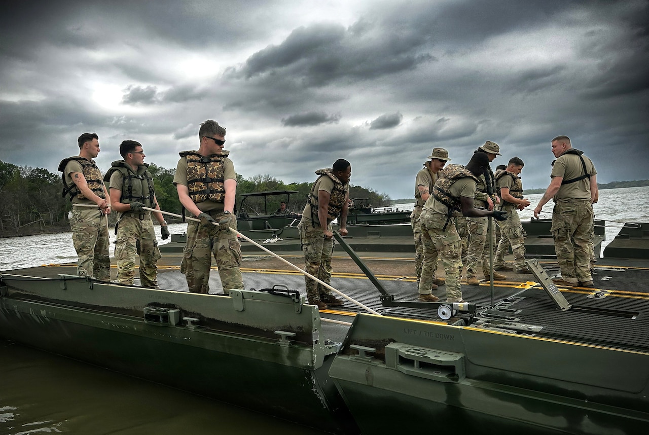 A dozen people in camouflage military uniforms and life jackets use ropes and other equipment to attach two sections of a floating bridge.