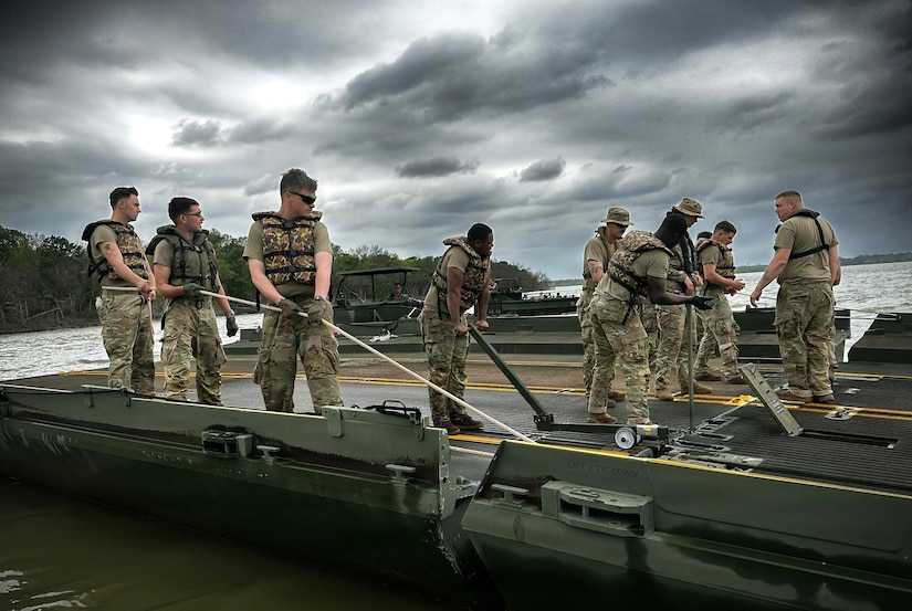 A dozen people in camouflage military uniforms and life jackets use ropes and other equipment to attach two sections of a floating bridge.