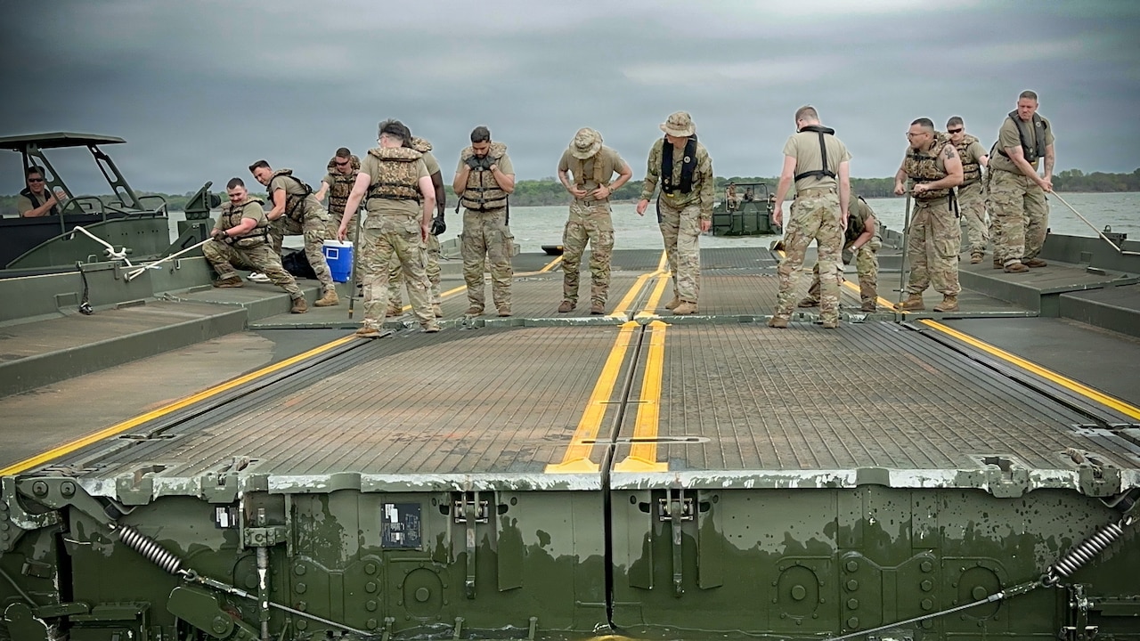 About a dozen people in camouflage military uniforms and life jackets attach two sections of a floating bridge while on a lake.