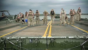 About a dozen people in camouflage military uniforms and life jackets attach two sections of a floating bridge while on a lake.