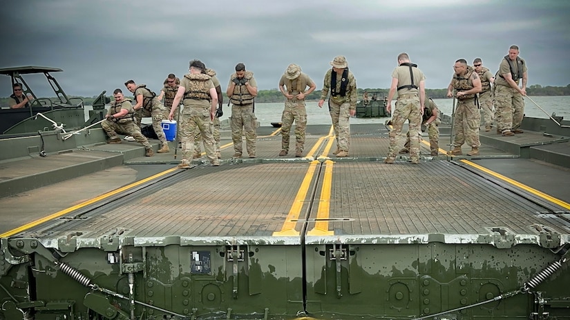 About a dozen people in camouflage military uniforms and life jackets attach two sections of a floating bridge while on a lake.