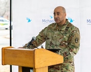 An Army colonel wearing combat uniform is speaking at a light brown podium.