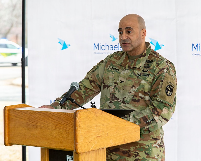 An Army colonel wearing combat uniform is speaking at a light brown podium.