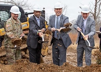 Three men in business suits (on right) and one man in Army combat uniform are turning soil with shovels in the air. All are wearing white construction helmets, and behind them is yellow and black construction equipment.
