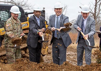 Three men in business suits (on right) and one man in Army combat uniform are turning soil with shovels in the air. All are wearing white construction helmets, and behind them is yellow and black construction equipment.