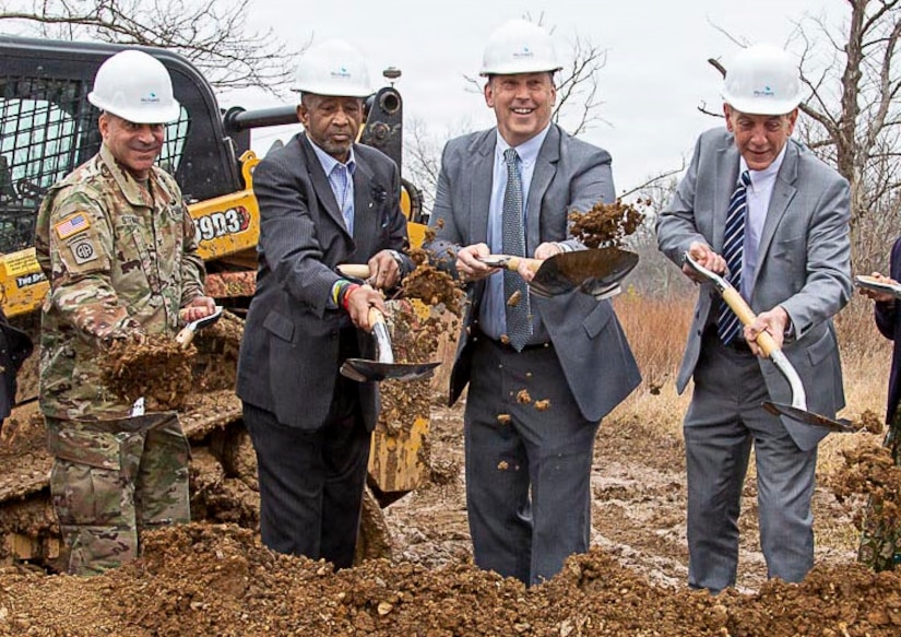 Three men in business suits (on right) and one man in Army combat uniform are turning soil with shovels in the air. All are wearing white construction helmets, and behind them is yellow and black construction equipment.
