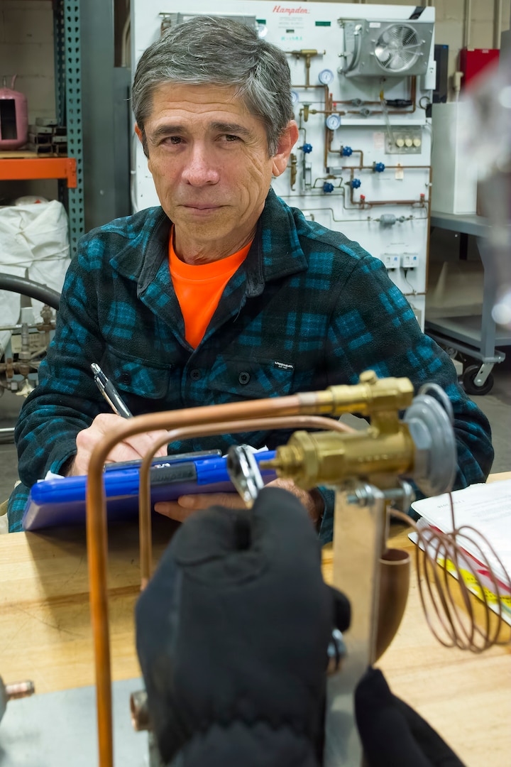 Donald Thompson, an air conditioning equipment mechanic with Pearl Harbor Naval Shipyard and Intermediate Maintenance Facility, observes another mechanic demonstrate how to adjust a refrigeration thermal expansion valve on a training device, Feb. 24, 2026. Thompson assesses mechanics to ensure they can troubleshoot and not just replace parts. PHNSY & IMF’s mission is to keep the Navy’s fleet “Fit to Fight” by repairing, maintaining, and modernizing the Navy's fast-attack submarines and surface ships. Strategically located in the heart of the Pacific, it is the most comprehensive fleet repair and maintenance facility between the U.S. West Coast and the Far East.