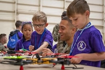 A service member kneels next to three kids in purple shirts as they touch drones sitting on a table.