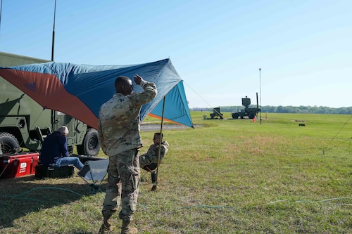 an Airmen shields his eyes from the sun's glare while looking to the sky. he's standing next to a tent where other service members are sitting